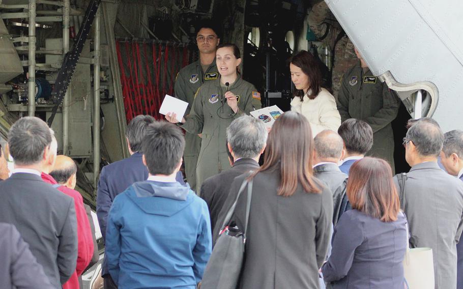 Two American service members in military flight suits stand in the open rear of a large cargo aircraft and speak to an assembled group of Japanese government officials, who are seen from behind.