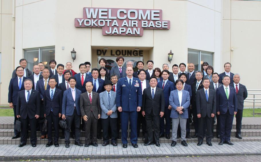 An American Air Force officer in blue dress uniform and a large group of Japanese government officials pose in suits on the steps outside a tan-colored building, with a sign reading “Welcome Yokota Air Base Japan” on the building above their heads.