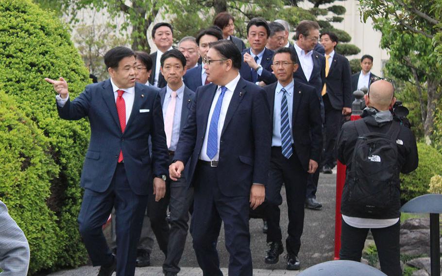 Several Japanese government officials in suits walk along a paved path with trees and bushes on either side during a tour of a military base.