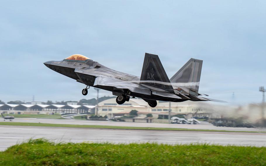 U.S. Air Force F-22 Raptor fighter jet takes off from the runway at Kadena Air Base in Okinawa, with landing gear retracting and base buildings visible in the background.