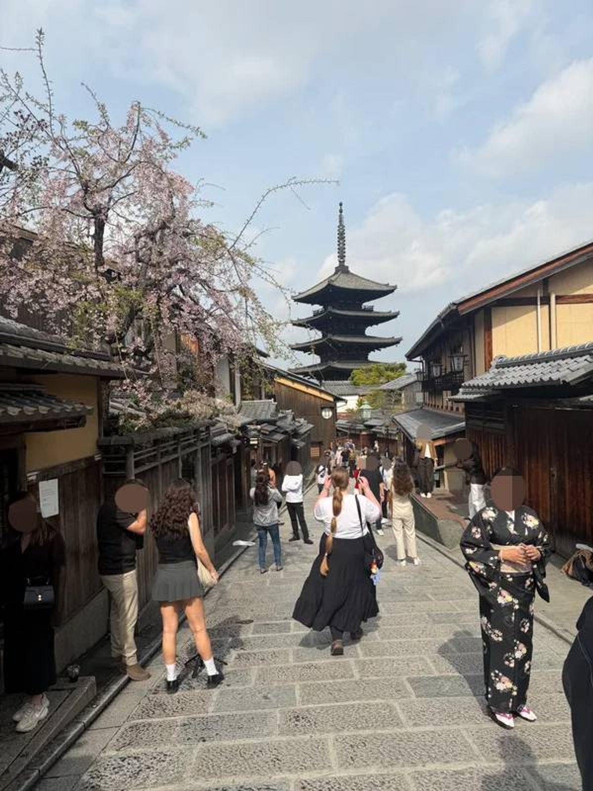 An image of a bustling street lined with traditional buildings, adorned with vibrant cherry blossoms. In the background, a prominent pagoda stands tall, drawing the eye amidst the activity of numerous pedestrians strolling leisurely.