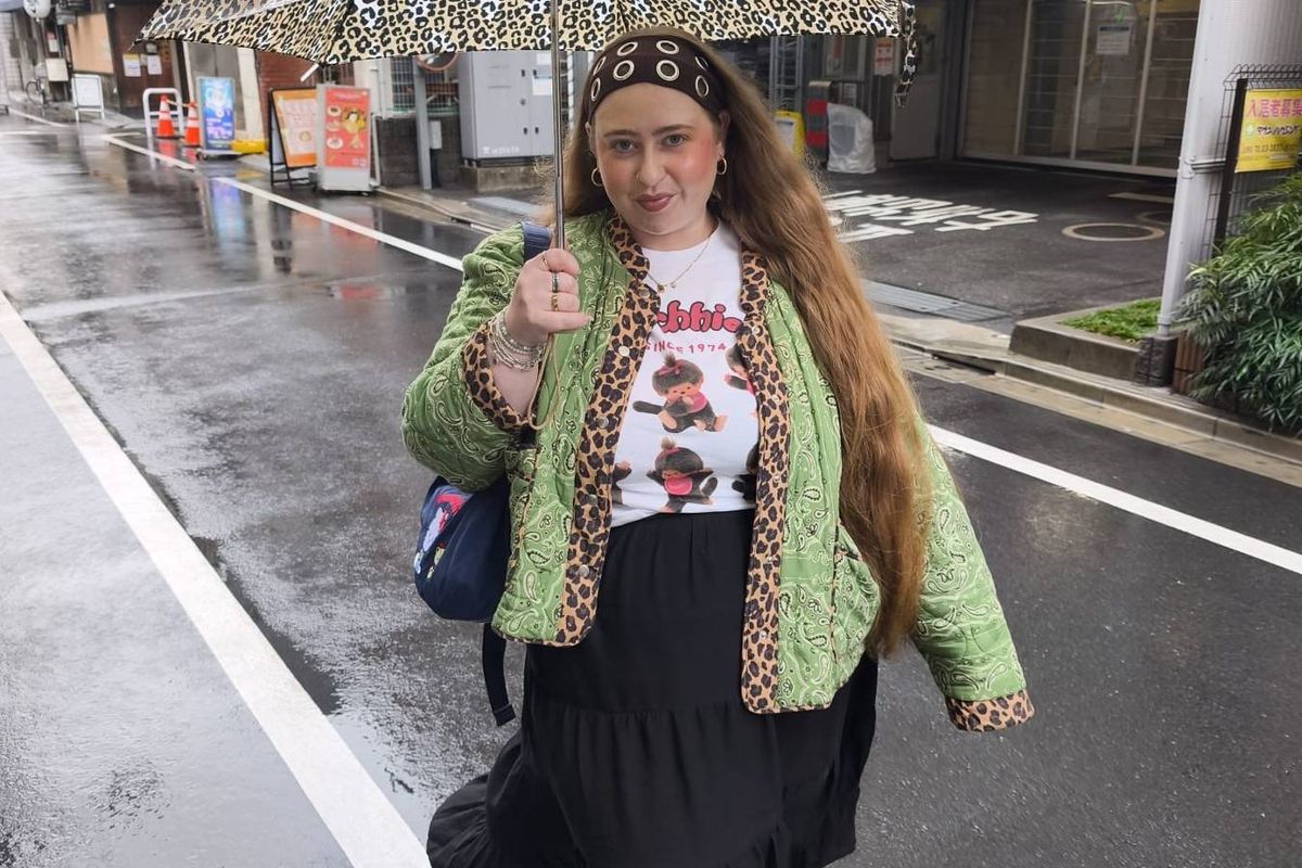 An individual, dressed in a green jacket and a patterned shirt, stands on a wet street during a rainfall. The person holds an umbrella in one hand and a handbag in the other, with several buildings visible in the background.
