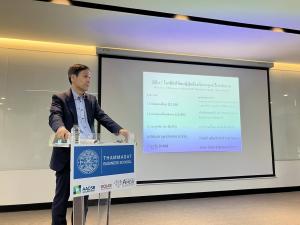 Dr. Koji Kurusu standing at a clear acrylic lectern marked "Thammasat Business School" (with AACSB, EQUIS, and AMBA accreditation badges), presenting a slide in Thai titled "Japan: Top 5 conditions leading to dependency on care" — dementia 12.3%, cerebrov