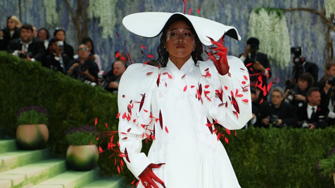 a woman attends the met gala in a long, white coat and a white hat with red feathers
