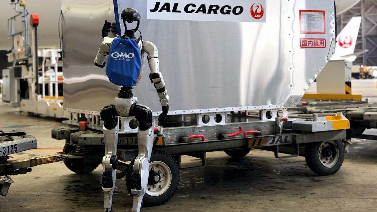 A humanoid robot stands near a baggage cart.