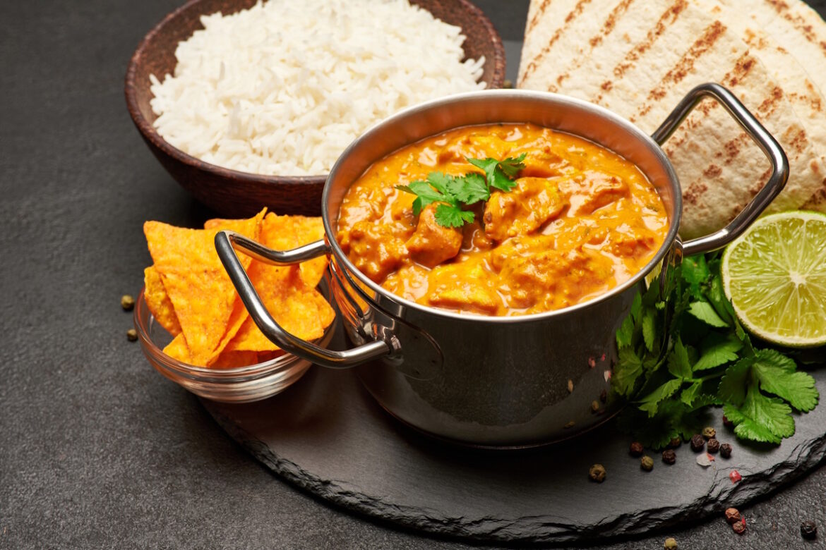 Pot of Traditional Chicken Curry, bowl of rice and pita on stone serving board.