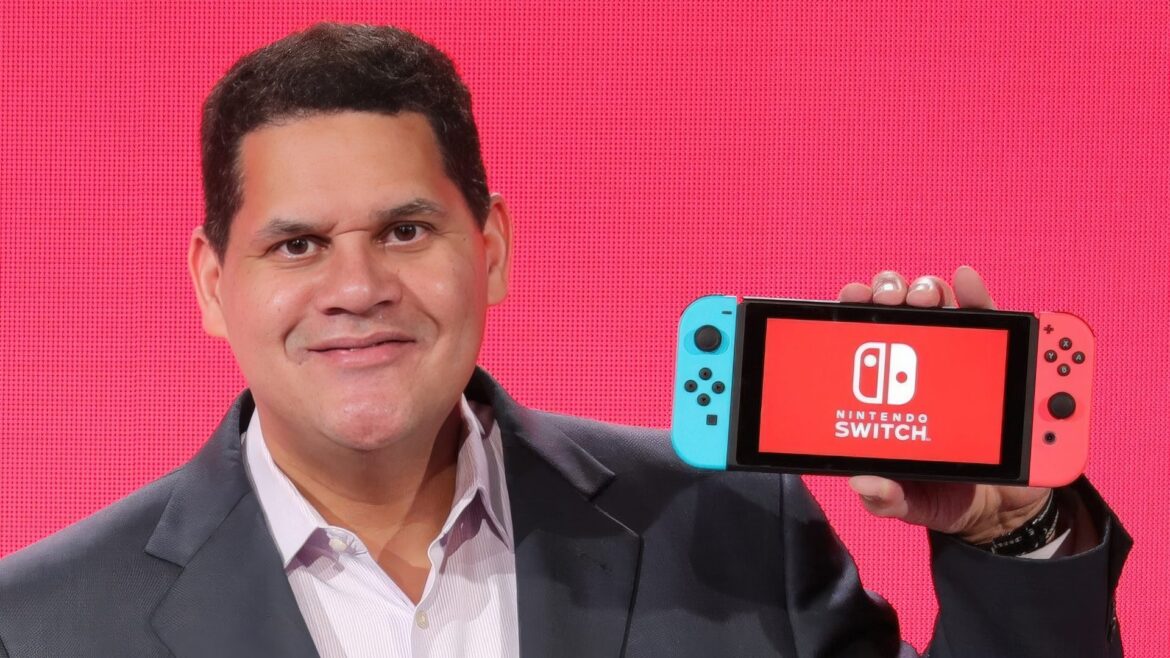 Reggie Fils-Aimé Defends Nintendo’s Full-Price Forever Policy, Pointing to Kyoto Roots Most Gamers Never Consider A person holding a Nintendo Switch with the Nintendo logo on the screen against a red background.