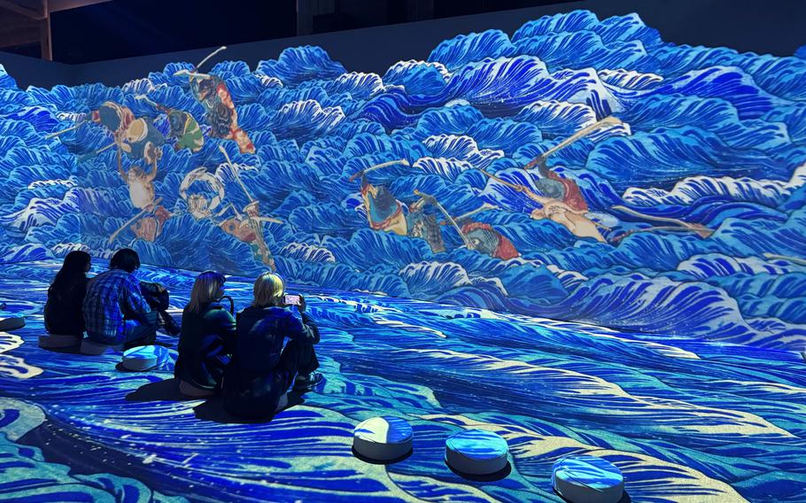 Projections of waves roll across the walls and floor as people sit on stools and watch.
