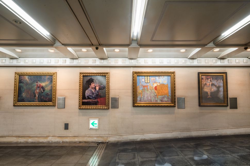 An underground gallery wall displays four framed reproductions of famous paintings with wall labels, lit by fluorescent ceiling lights above a tiled walkway.
