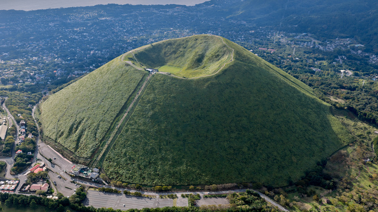 A volcano's crater covered in grass with a cityscape in the background