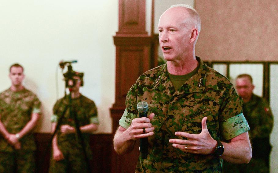 A Marine Corps officer in camouflage uniform speaks into a microphone while standing in a club ballroom, with other Marines in camouflage uniform stand against a wall behind him.