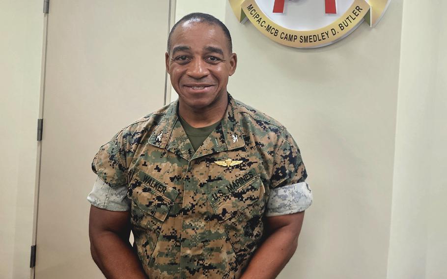 A Marine Corps officer in camouflage uniform poses in an office in front of a white wall, with a battalion emblem hanging above his left shoulder.