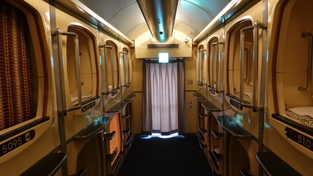 Corridor lined with classic cream-colored capsule hotel pods stacked two tiers high, with ladders to the upper units