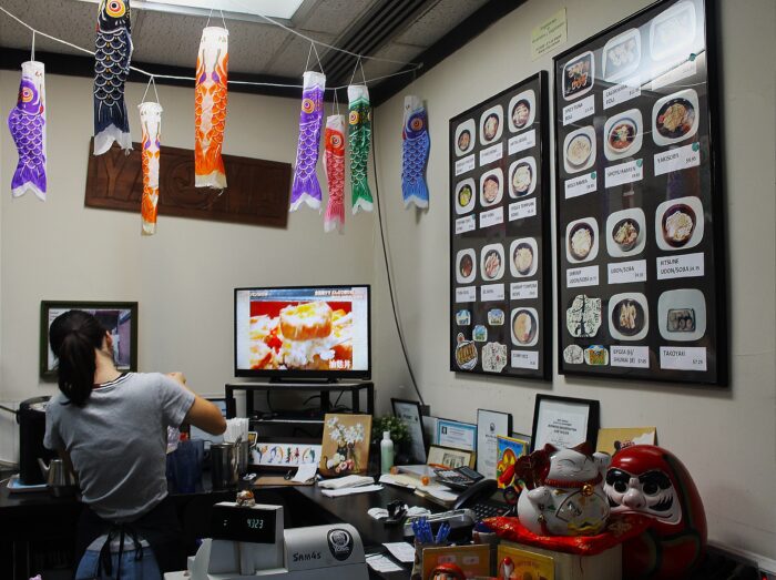 A woman stands at a counter while a cooking show is seen on the TV next to her. Above her are colorful fish decorations that look like they are made from paper.