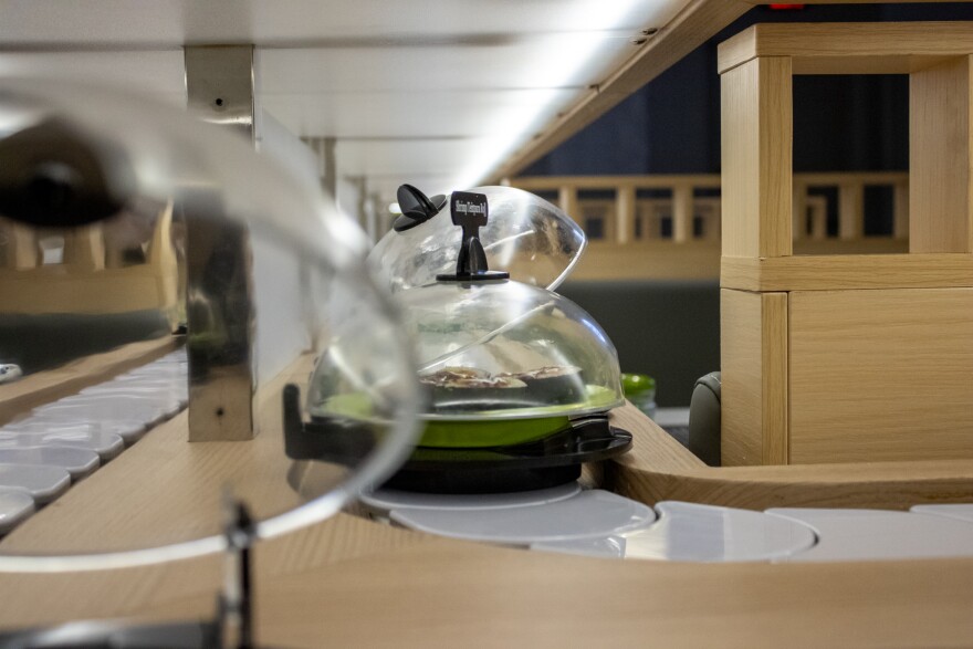 Small plates line a conveyor belt that wraps around a restaurant. Booths line either side. The image is taken from the perspective of the belt. It appears as if the camera is moving along and plates are coming toward it.