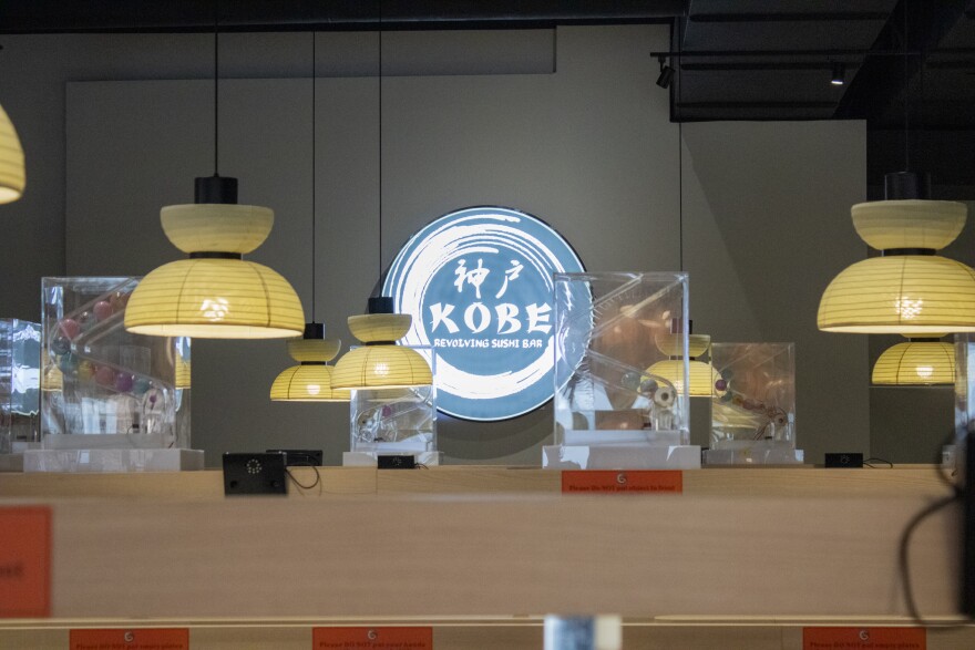 A sign for Kobe Revolving Sushi Bar on a wall. Light fixtures hang from the ceiling and contraptions that release small toys line a high shelf that wraps around the restaurant.