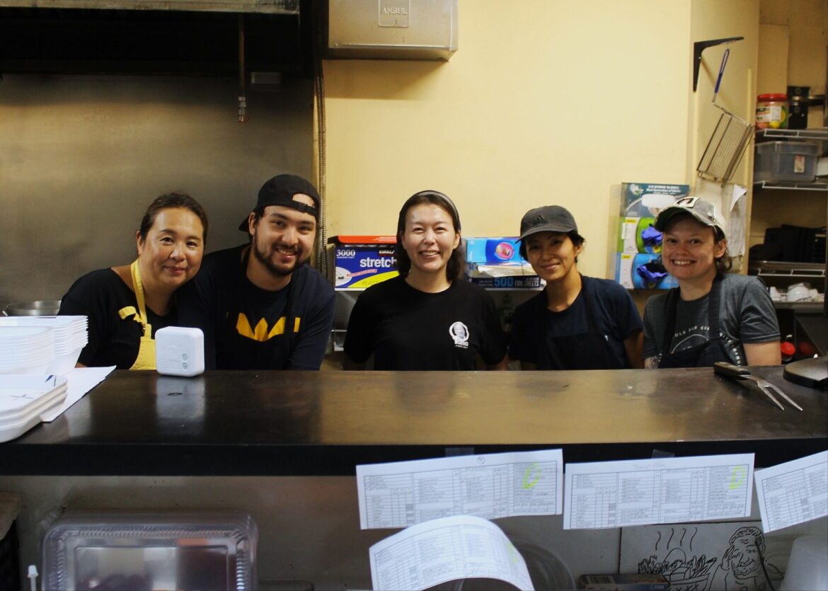 Yama: A Japanese Community Space In Morgantown, W.Va. Six smiling people are shown - five women and one man. They pose for a photo from behind a counter at a restaurant they work at. Some are dressed with black aprons.
