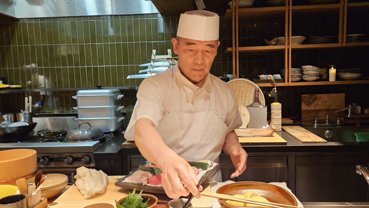 A man wearing a paper chef's hat, a white shirt and apron organizes fish, fruit and vegetables on a preparation counter