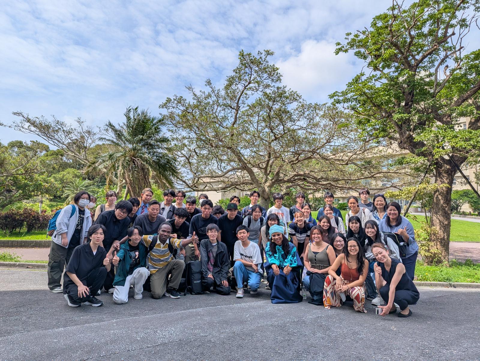 Group photo of a large diverse crowd posing outdoors in a sunny park with trees.