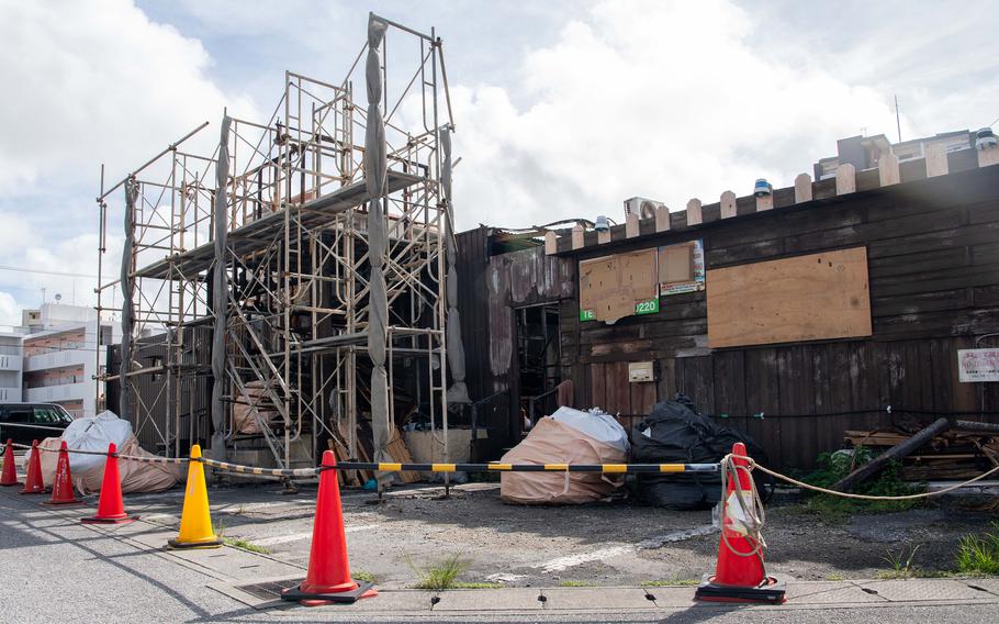 A fire-damaged building stands behind safety cones and caution tape as scaffolding surrounds part of the structure. Boarded windows, charred exterior walls and debris bags are visible outside the building.