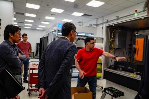 A man in a red shirt demonstrates an advanced manufacturing machine to a delegation of people in business wear.