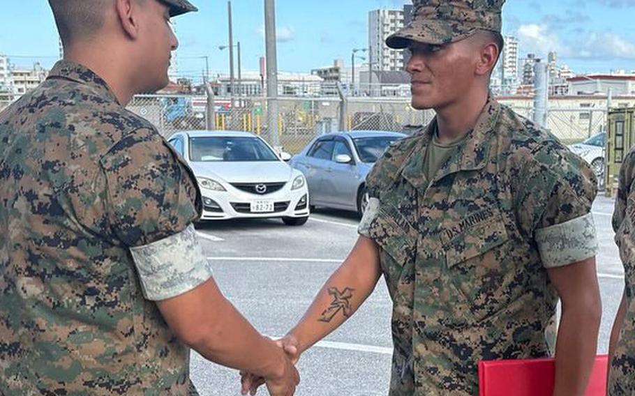 Two U.S. Marines in camouflage utility uniforms shake hands in a paved parking area during a military recognition or promotion event.