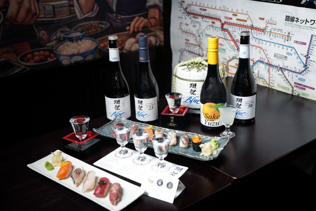 Four bottles of Dassai Blue sake displayed behind plates of sushi and small glasses of sake.