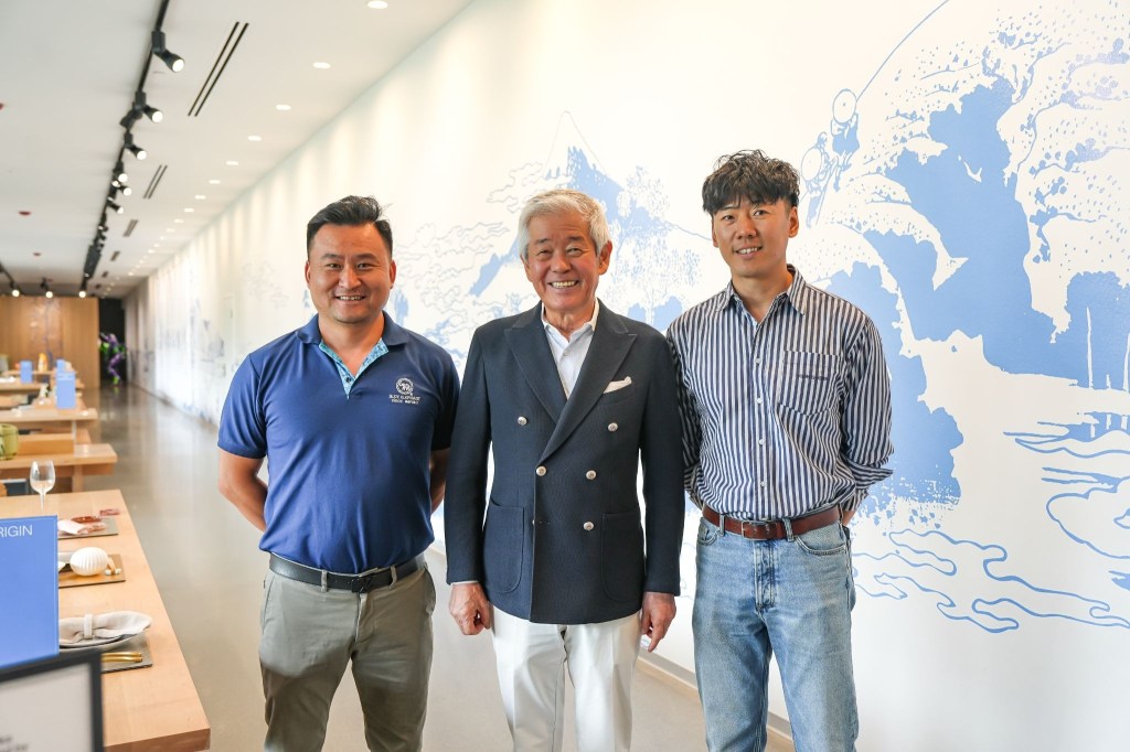Three men stand smiling together in an indoor space with a large blue and white wall mural behind them.